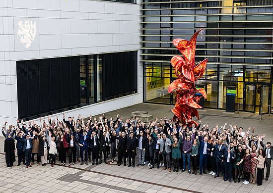 Übersichtsbild des Abschlussjahrgangs 2023/2024, der vor einer roten Skulptur vor Gebäude VW der BUW steht. Viele Personen lächeln und winken in die Kamera.