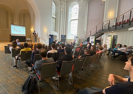 Besucherinnen und Besucher sitzen auf Stühlen inmitten des Raumes und hören einem Vortrag zu.