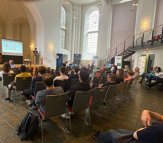 Besucherinnen und Besucher sitzen auf Stühlen inmitten des Raumes und hören einem Vortrag zu.