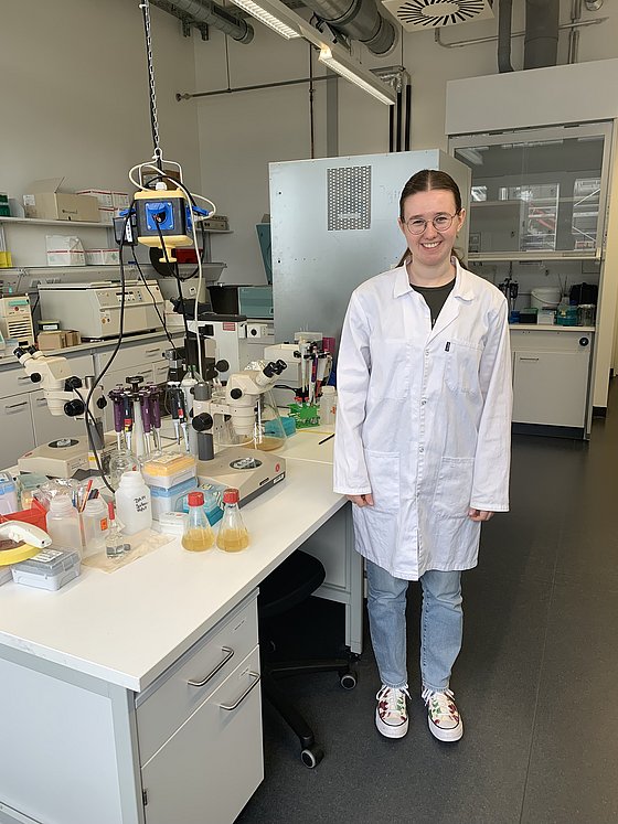 Intern Ida is standing at a lab bench with various utensils and a microscope, wearing a white lab coat and smiling at the camera.