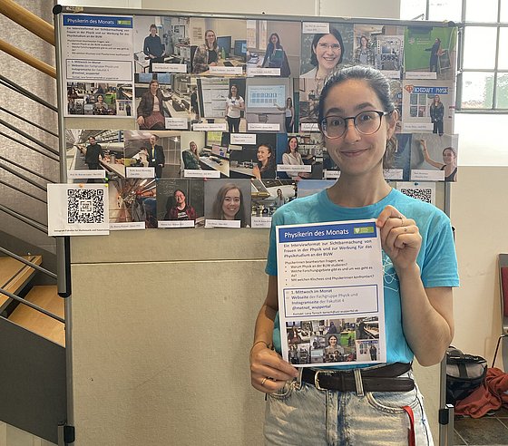 Eine Frau steht vor einer Foto-Collage und hält ein Info-Blatt zum Projekt "Physikerin des Monats" hoch.