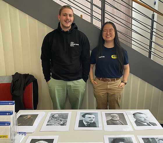 Eine Studentin und ein Student stehen an einem Tisch, auf dem schwarz-weiß Bilder von Physikerinnen und Physikern und Flyer liegen.