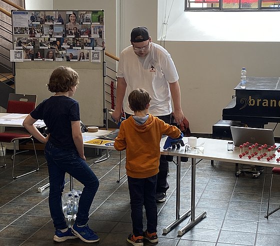 Zwei Kinder und ein Physikstudent stehen an einem Tisch mit einem Ausstellungsstück.